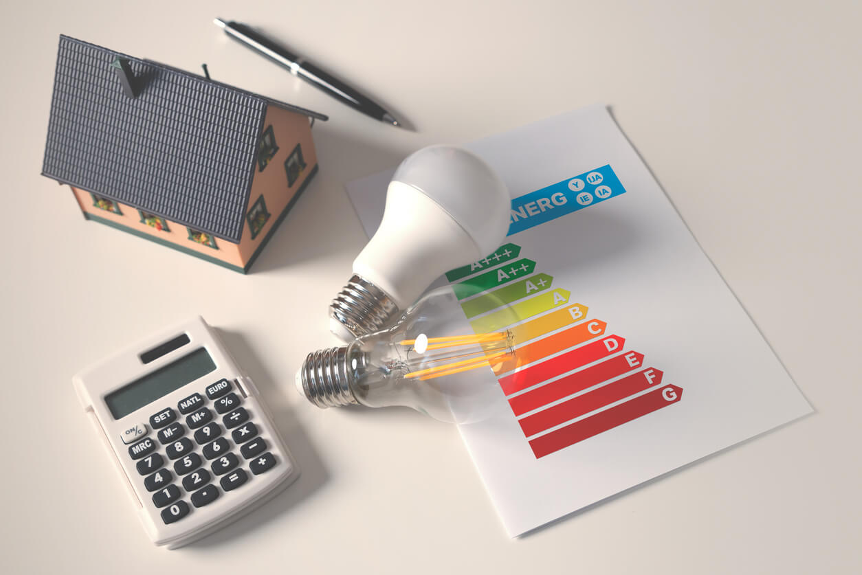 Lâmpadas, calculadora e tabela de eficiência energética ao lado de uma miniatura de casa, simbolizando o cálculo de consumo de energia em kWh.