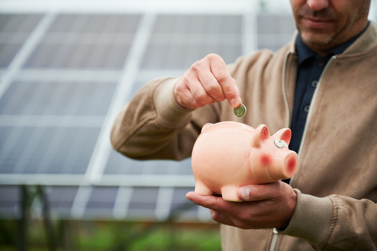 Homem colocando moeda em cofrinho, com painéis solares ao fundo, simbolizando economia e financiamento de energia solar.