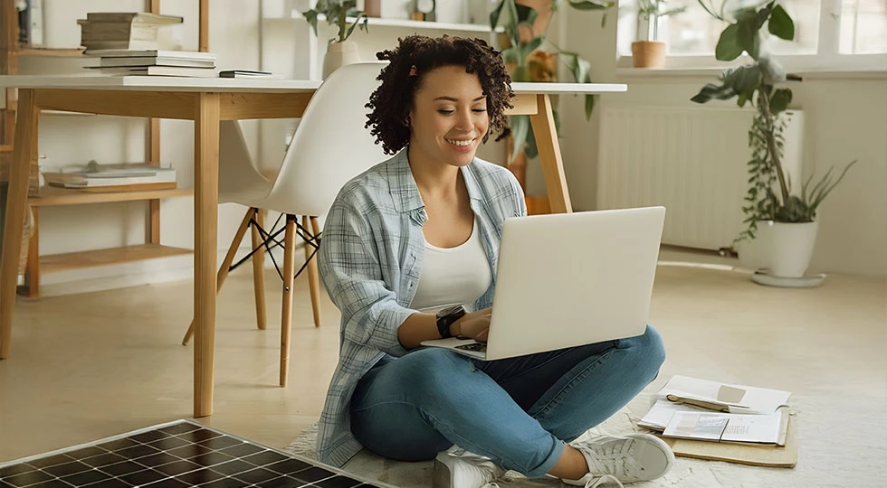 Mulher jovem sorridente usando laptop portátil em um ambiente bem iluminado, com plantas e móveis de madeira, criando um espaço de trabalho agradável.