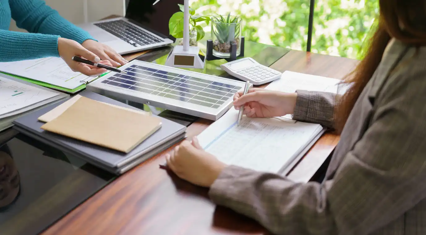 Pessoa analisando planos de energia solar em reunião de negócios, com diversas ferramentas de trabalho e plantas ao redor. Ambiente corporativo bem iluminado.