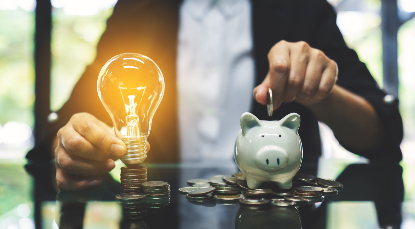 Mãos segurando uma lâmpada acesa ao lado de um cofrinho de porquinho, com moedas ao redor, simbolizando dicas práticas para economizar energia elétrica e reduzir custos.