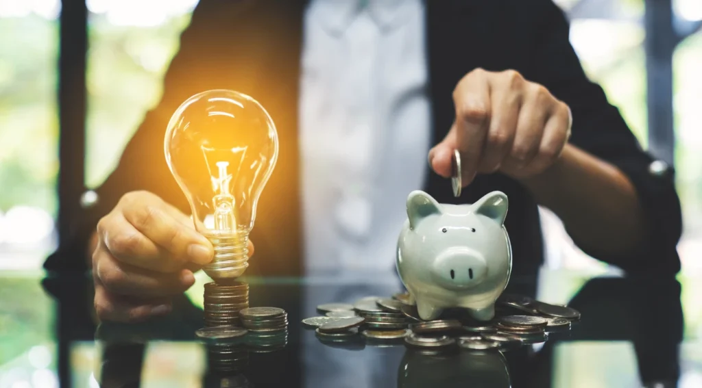 Pessoa colocando uma moeda na caixa de porquinho ao lado de uma lâmpada de ideia acesa, simbolizando economia e inovação financeira.