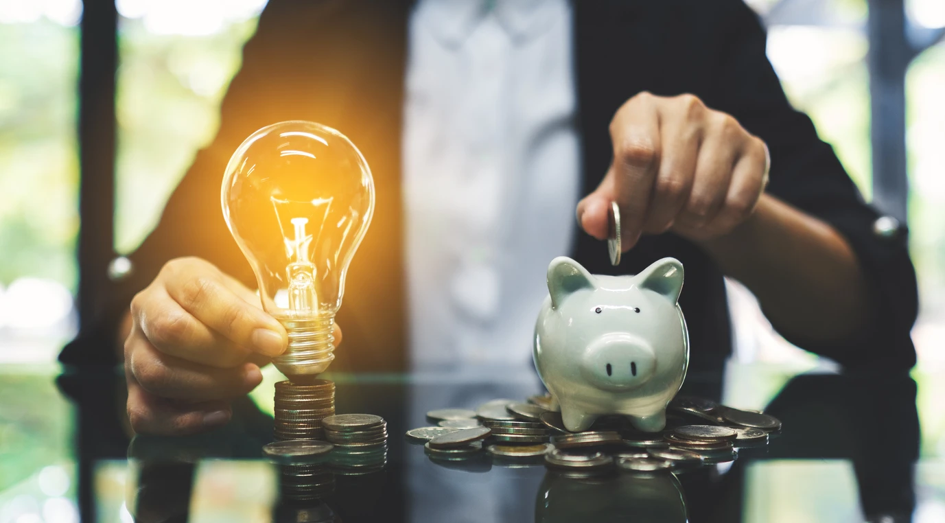 Pessoa colocando uma moeda dentro de um cofrinho de porquinho de cerâmica ao lado de uma lâmpada de led acesa, representando economia de energia ou investimento financeiro.