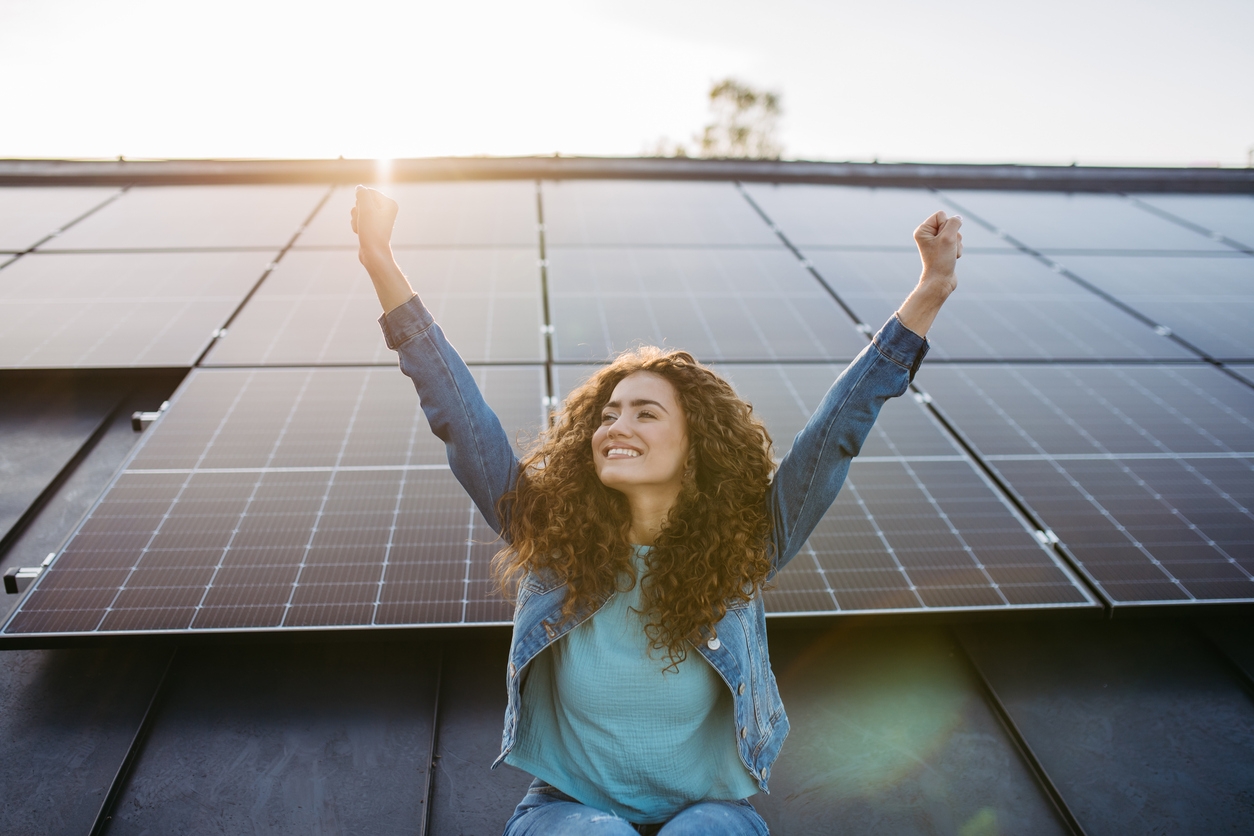 Mulher sorridente com os braços erguidos em celebração, sentada em frente a painéis solares, representando a conquista de energia sustentável e economia com energia solar.