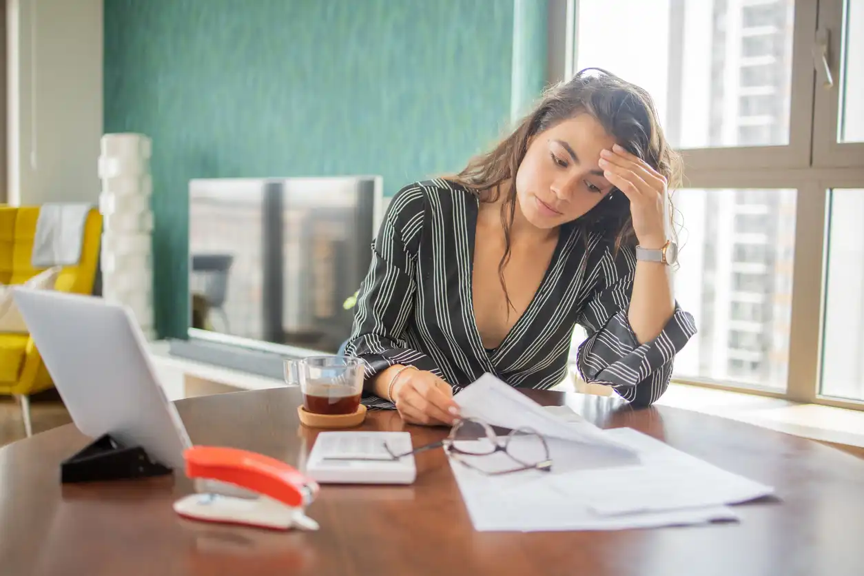 Mulher com expressão preocupada analisando documentos em um escritório com janela grande ao fundo, ambiente de trabalho moderno.