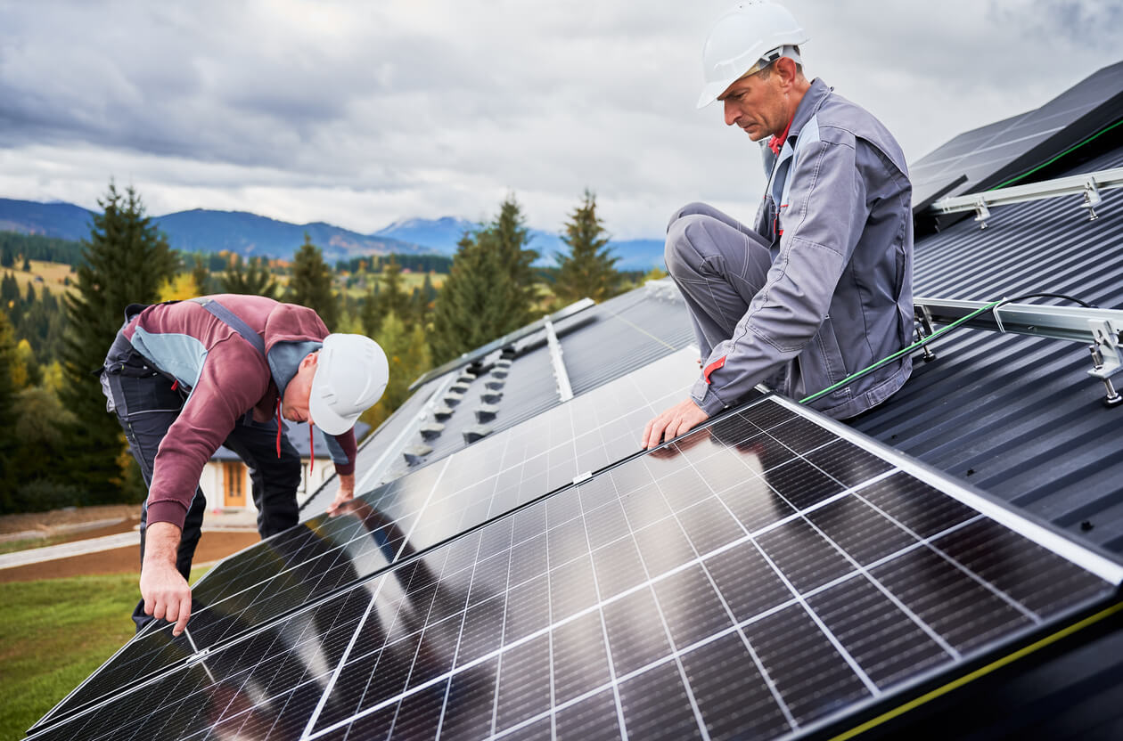 Dois técnicos instalando painéis solares em um telhado em meio a uma paisagem montanhosa, representando a adoção de energia solar como uma solução sustentável. Imagem ideal para explorar se a energia solar vale a pena, destacando benefícios como economia e preservação ambiental.