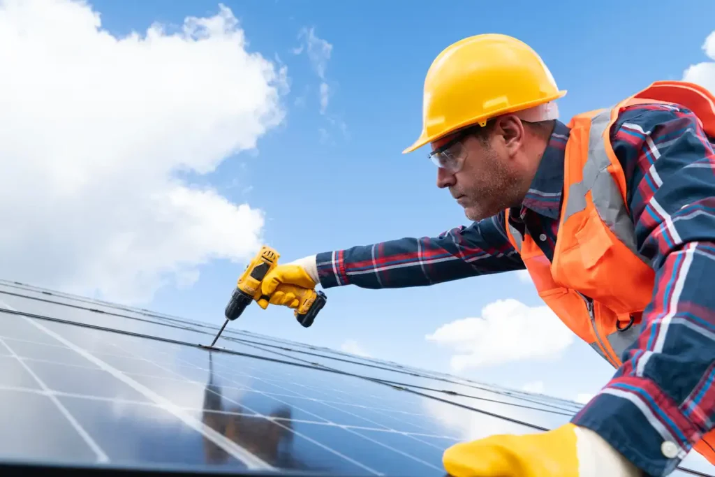 Eletricista instalando painéis solares ao ar livre ensolarado, usando capacete amarelo, vestimenta de segurança laranja e ferramentas elétricas.