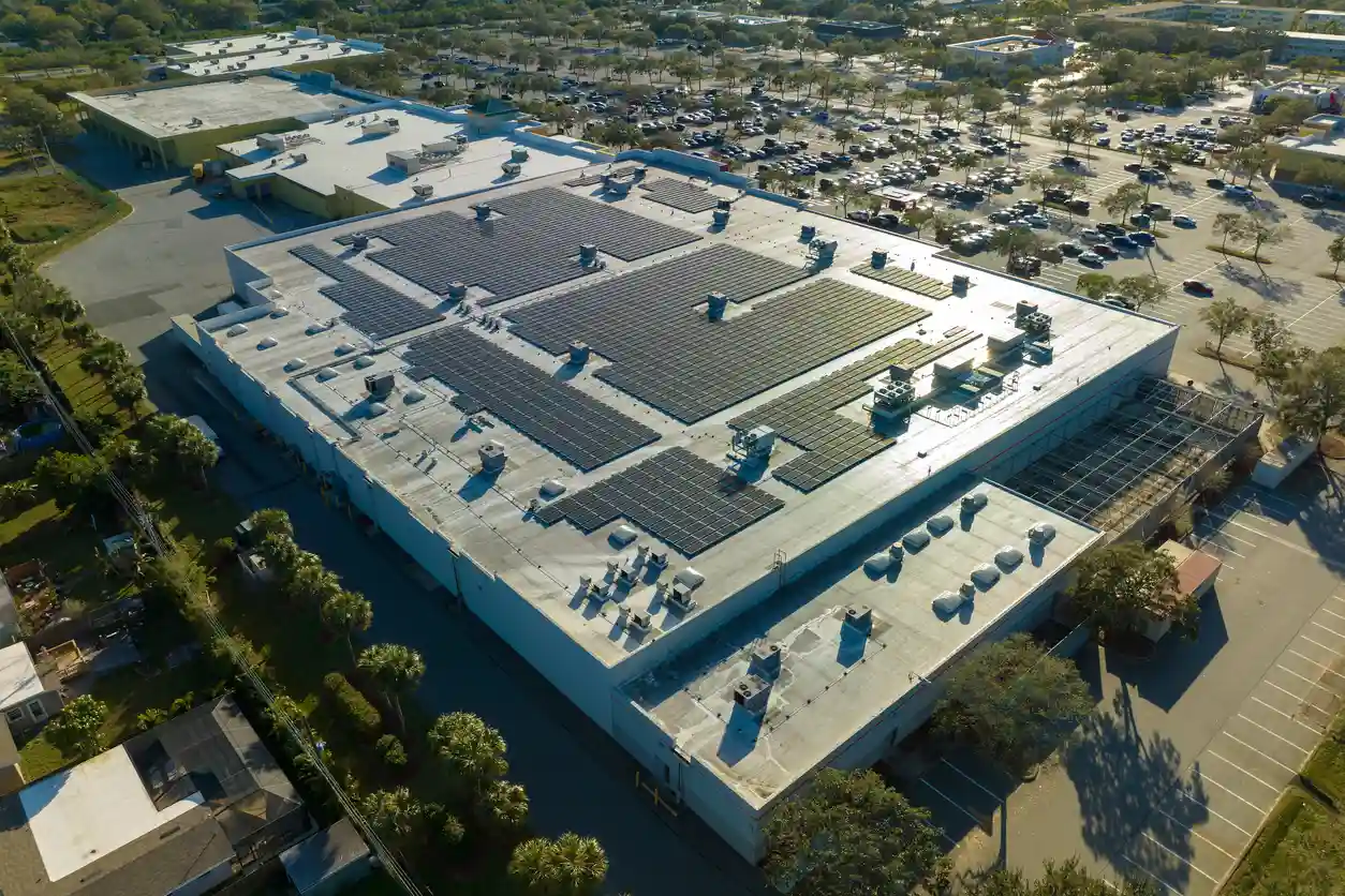 Imagem aérea de um grande supermercado com painéis solares instalados no telhado, localizado em uma área urbanizada com estacionamento e árvores ao redor.