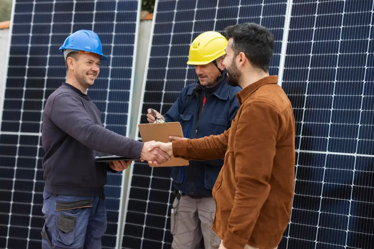 Três homens em uma usina solar trocando um aperto de mão, um deles com capacete azul, outro com capacete amarelo, e o terceiro com cabelo escuro, simbolizando energia renovável e sustentabilidade.