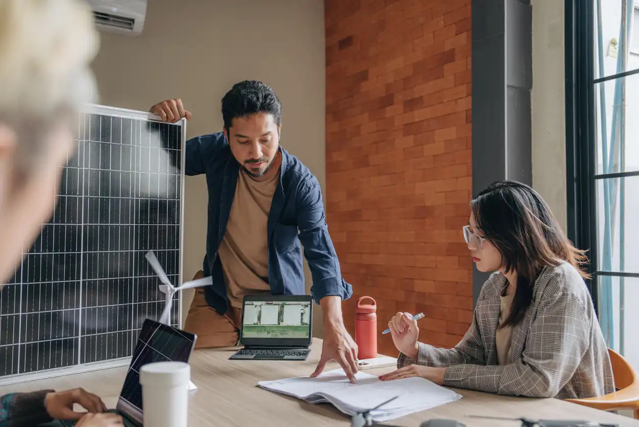 Engenheiros discutindo energia solar e energias renováveis em uma reunião de projeto, com painel solar e laptop na mesa.