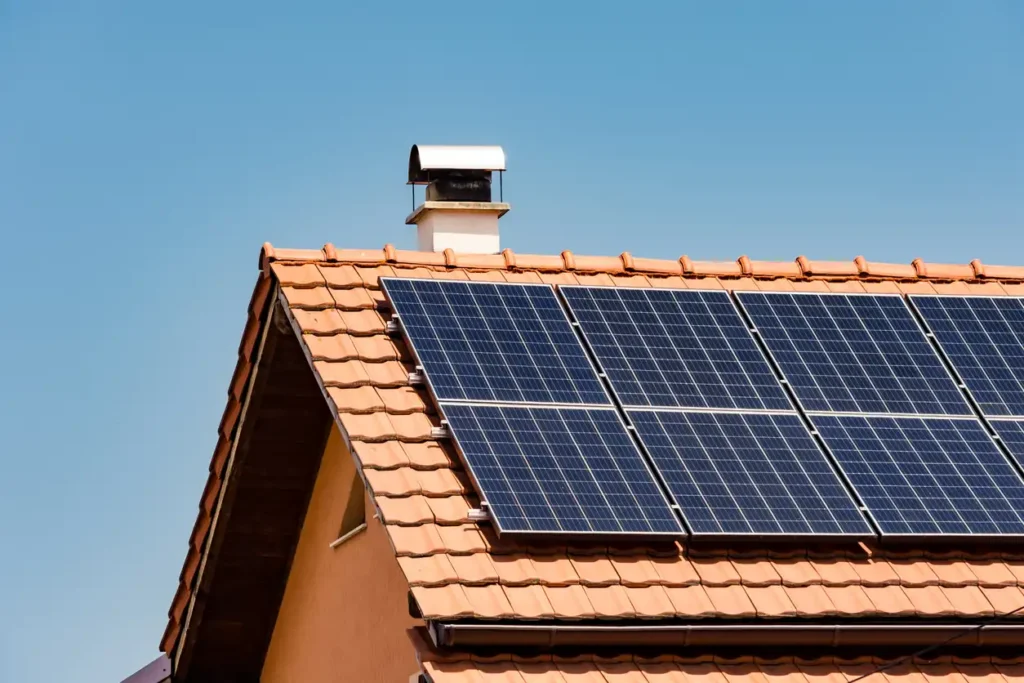 Painel solar instalado no telhado de uma casa com telhas de cerâmica, captando energia solar de um céu limpo e azul.