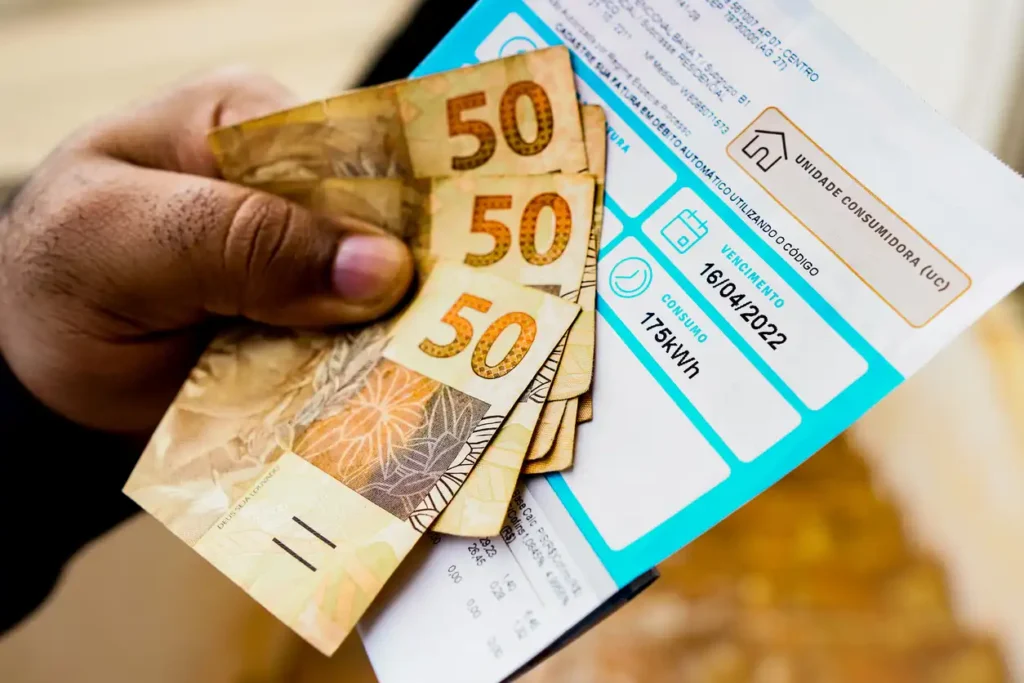 Pessoa segurando várias cédulas de cinquenta reais e uma conta de energia elétrica digital, ilustrando o pagamento de contas de energia com dinheiro.