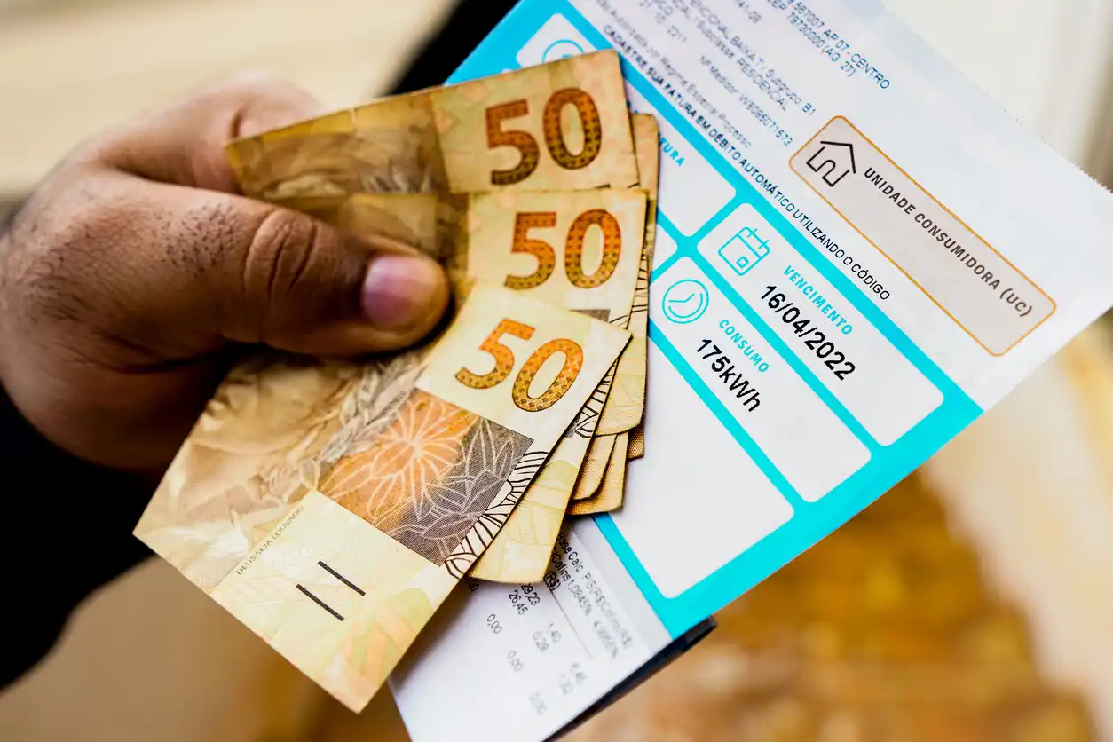 Pessoa segurando várias cédulas de cinquenta reais e uma conta de energia elétrica digital, ilustrando o pagamento de contas de energia com dinheiro.