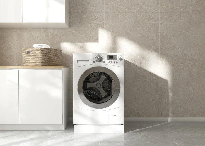 Lavanderia moderna com máquina de lavar roupa branca, bancada com cesto de roupa limpa, parede de azulejos bege e piso de porcelanato cinza, iluminação natural.