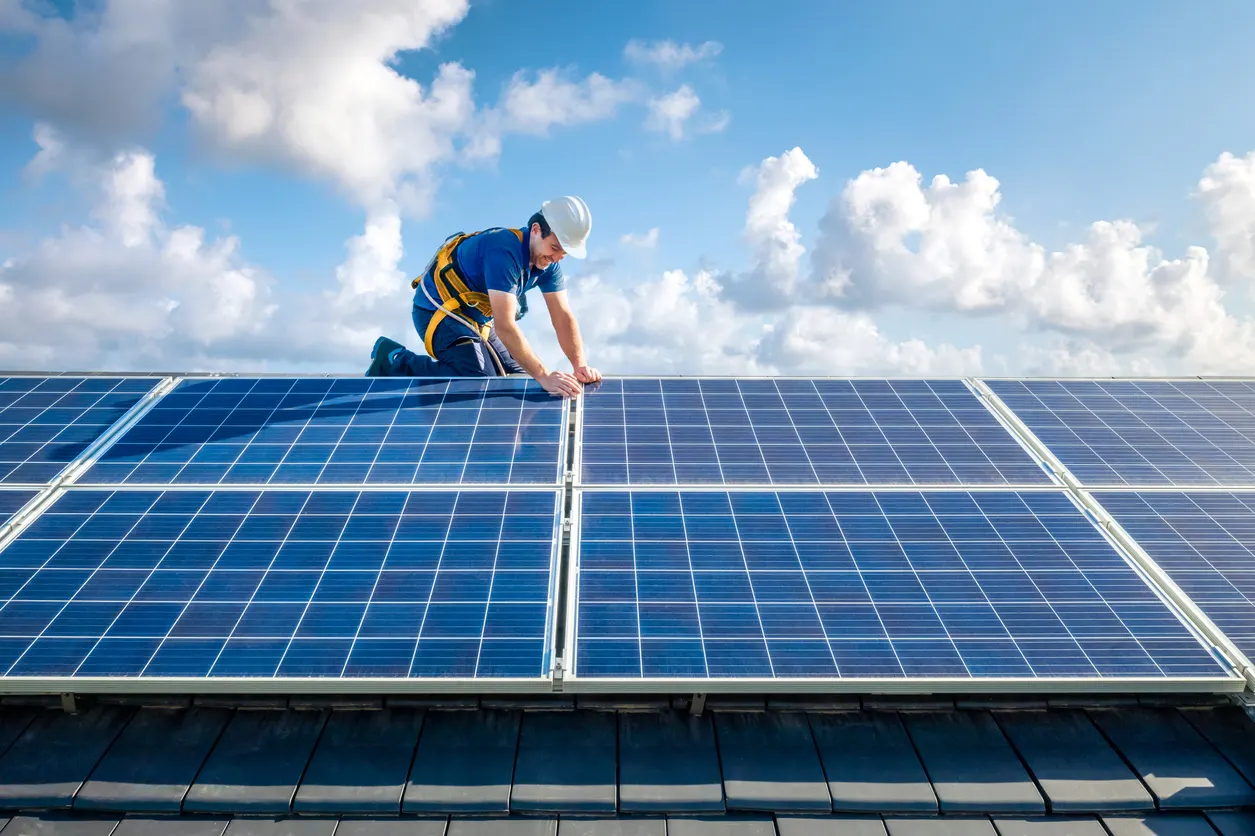 Técnico de energia solar instalado ajustando um painel fotovoltaico azul em uma usina solar no topo de um prédio, com céu azul e nuvens ao fundo.