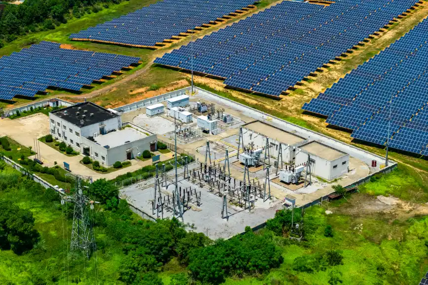 Vista aérea de uma usina solar com grandes painéis fotovoltaicos instalados em sequência e, ao centro, uma subestação elétrica com estruturas metálicas e edifícios de apoio.