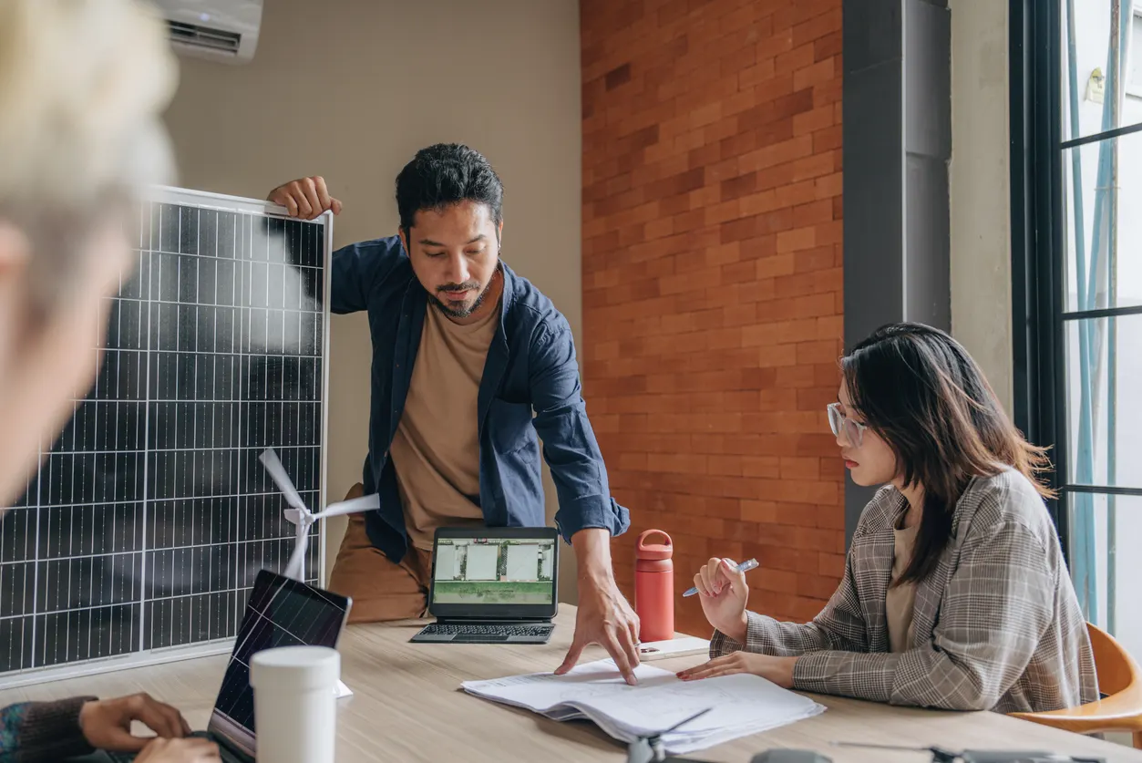 Equipe em escritório com placa solar ao fundo: homem em pé aponta dados em um notebook, enquanto mulher analisa documentos na mesa, em reunião técnica sobre energia e sustentabilidade.