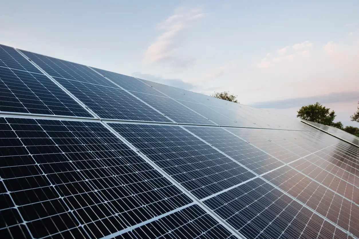 Painéis de energia solar fotovoltaica em grande escala, com células azuis e céu ao fundo ao entardecer, mostrando a estrutura de captação de luz.