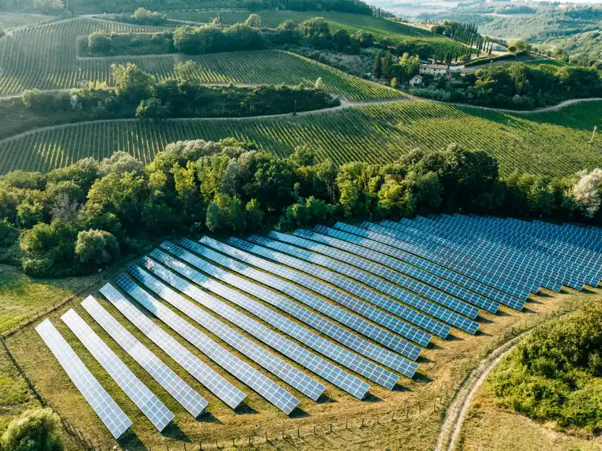 Vista aérea de um amplo campo com painéis solares fotovoltaicos instalados em meio a áreas rurais e colinas verdes, em um cenário de fazenda